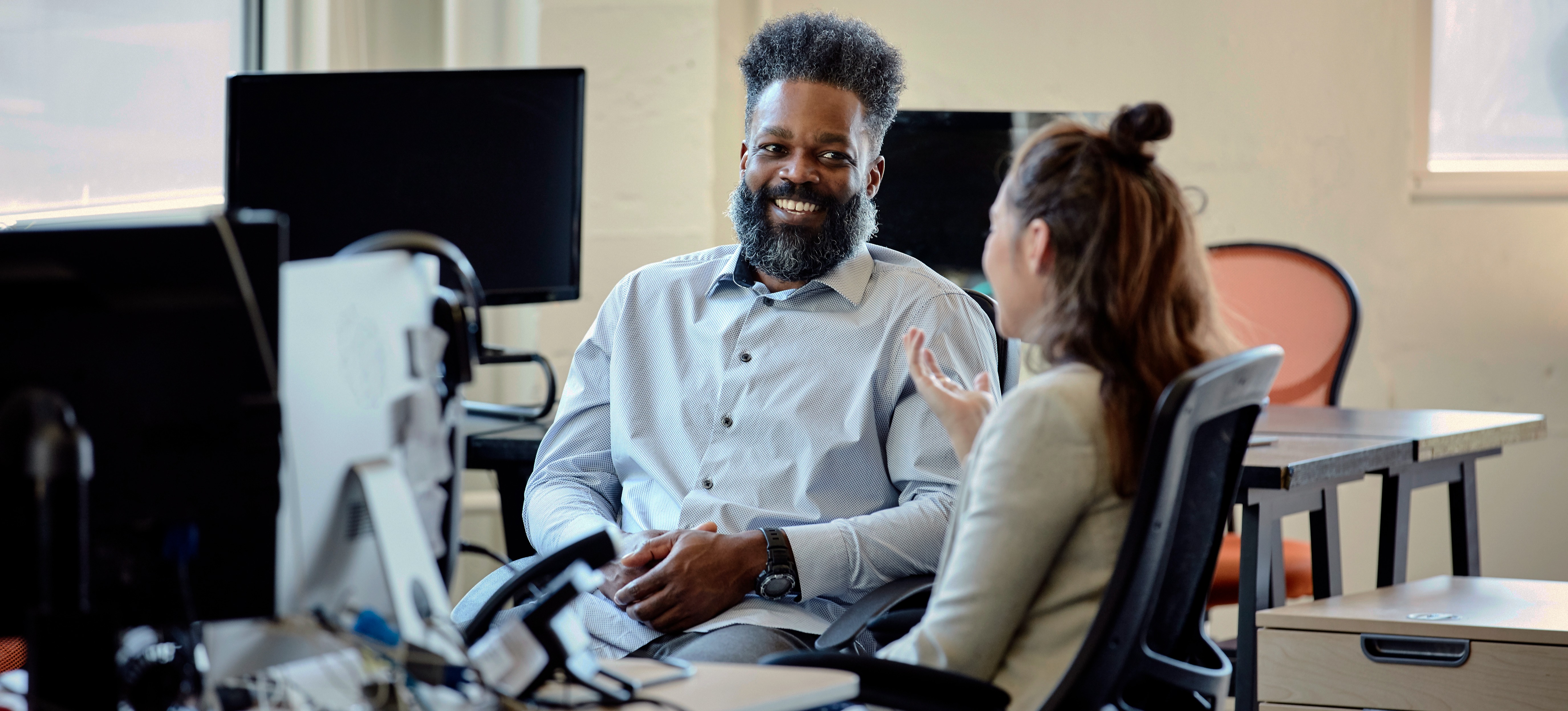 [Featured Image] A bearded mentor in a grey shirt speaks to a client with a hair bun and a grey suit jacket. 