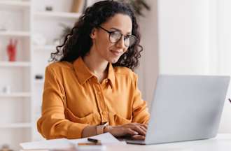 [Featured image] A woman in an orange shirt works on her laptop at a white table.