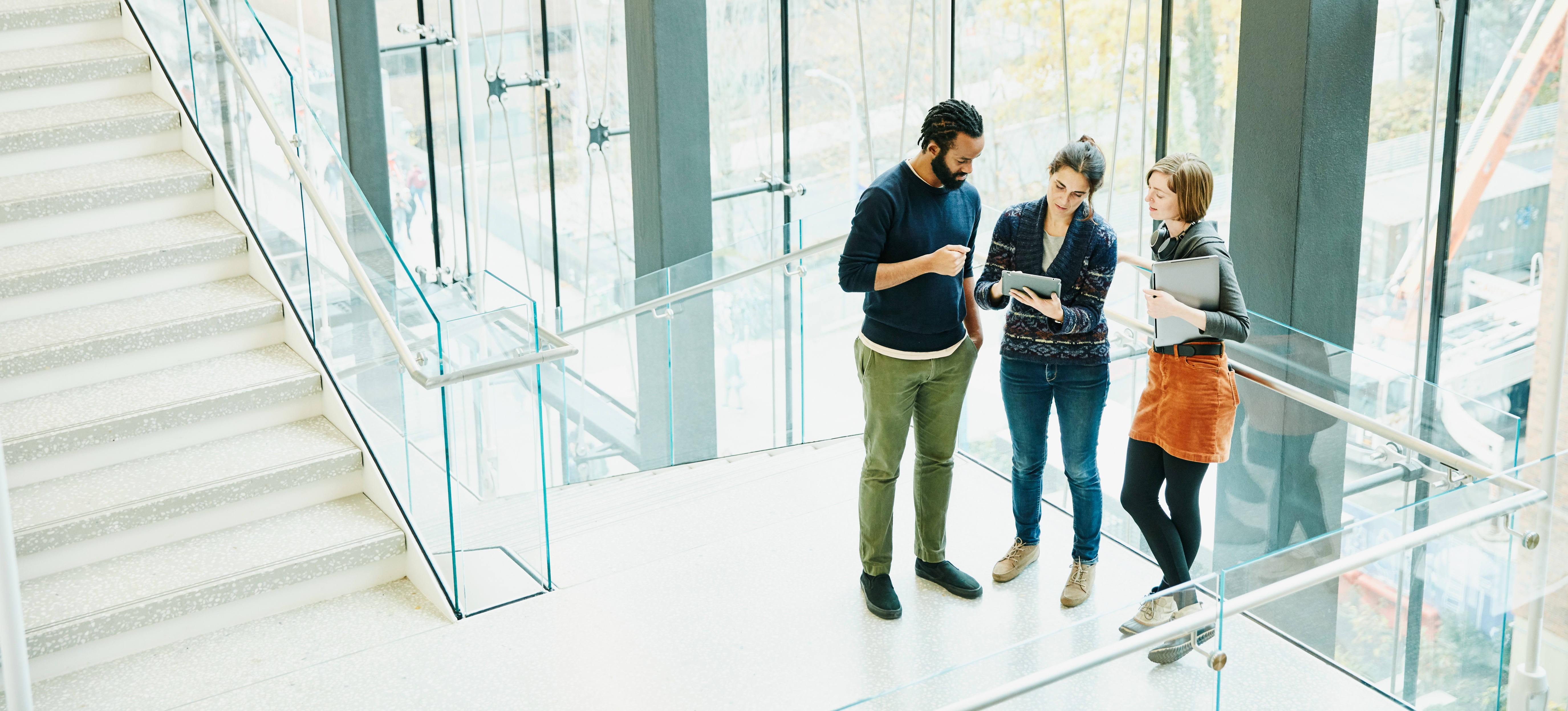 [Featured Image] Three coworkers stand in an office building reviewing information on a tablet. 