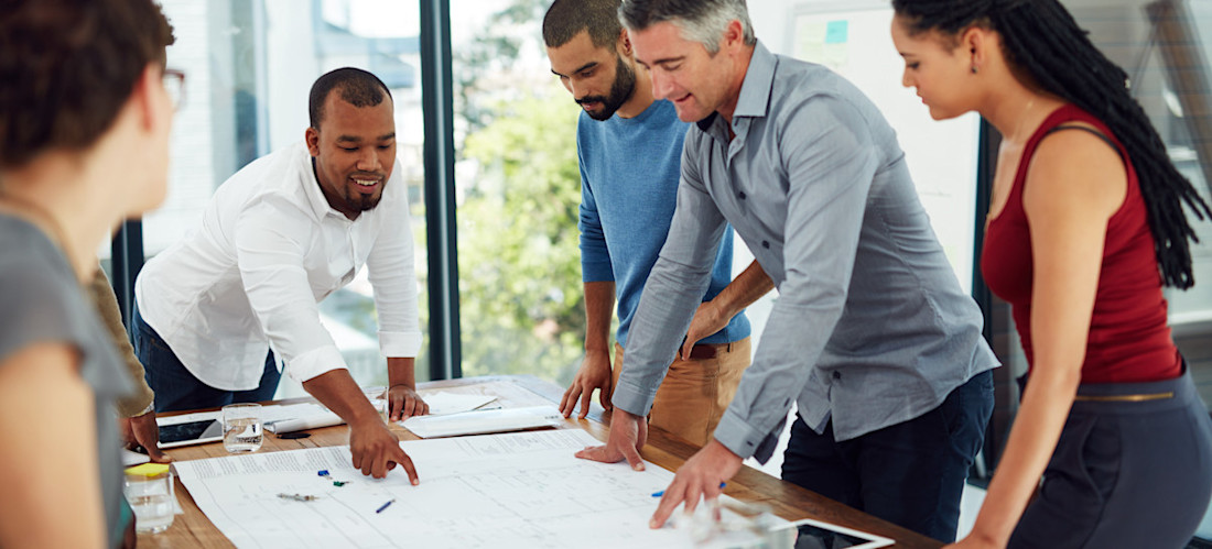 [Featured image] A Scrum product owner who has received CSPO certification stands at a table with their project team and discusses project plans.