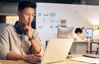 [Featured Image] A programmer sits at his desk and helps his company integrate AI.  