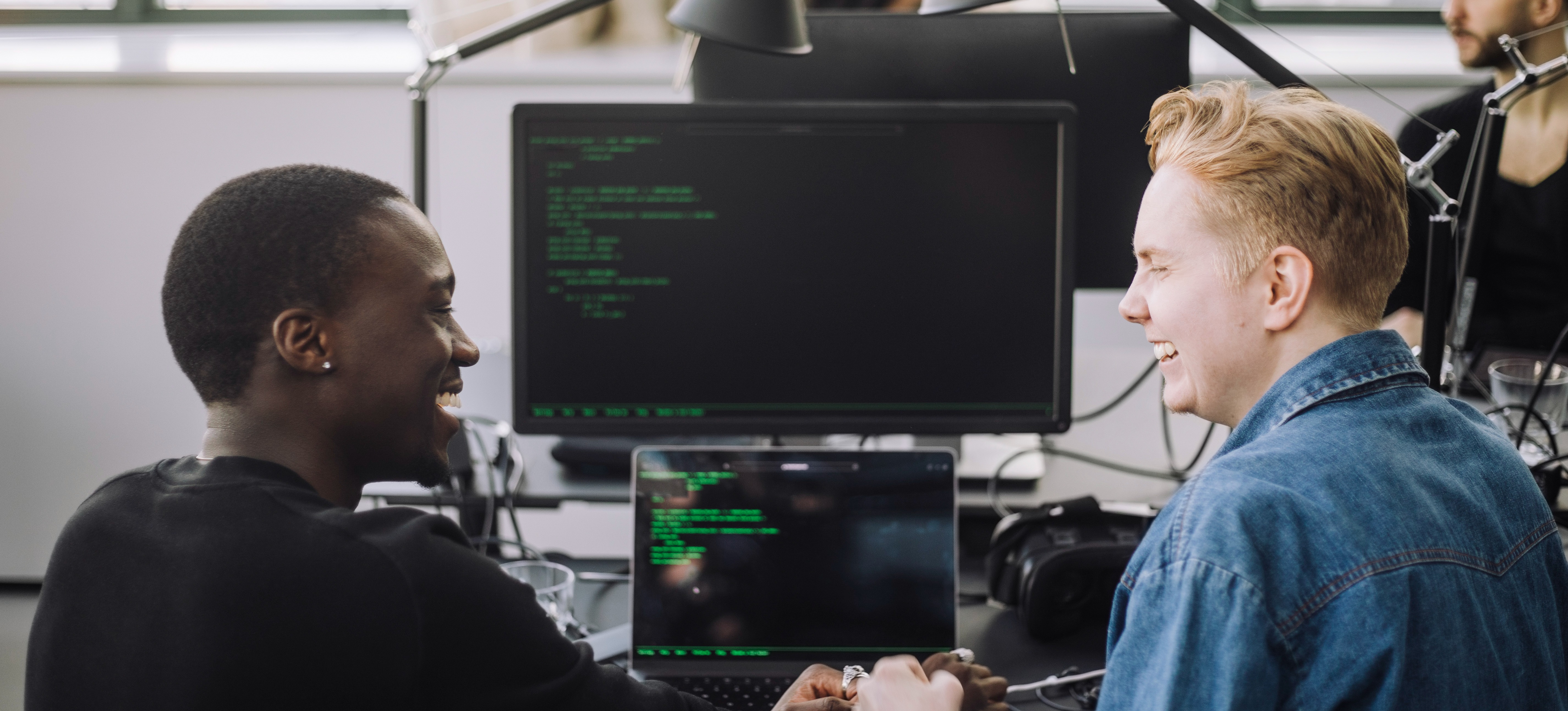 [Featured Image] Cybersecurity professionals in a work environment discussing cybersecurity tools while implementing security measures on their computers.
