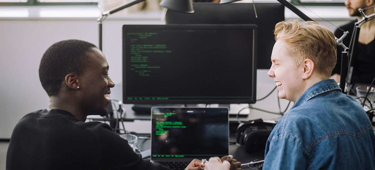 [Featured Image] Cybersecurity professionals in a work environment discussing cybersecurity tools while implementing security measures on their computers.
