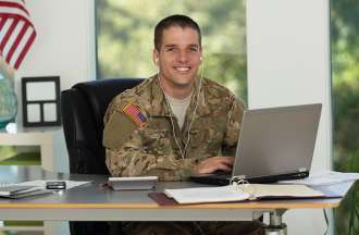 [Featured image] A cryptanalyst wearing army fatigues and white earphones works on a laptop at a desk in front of three windows. 