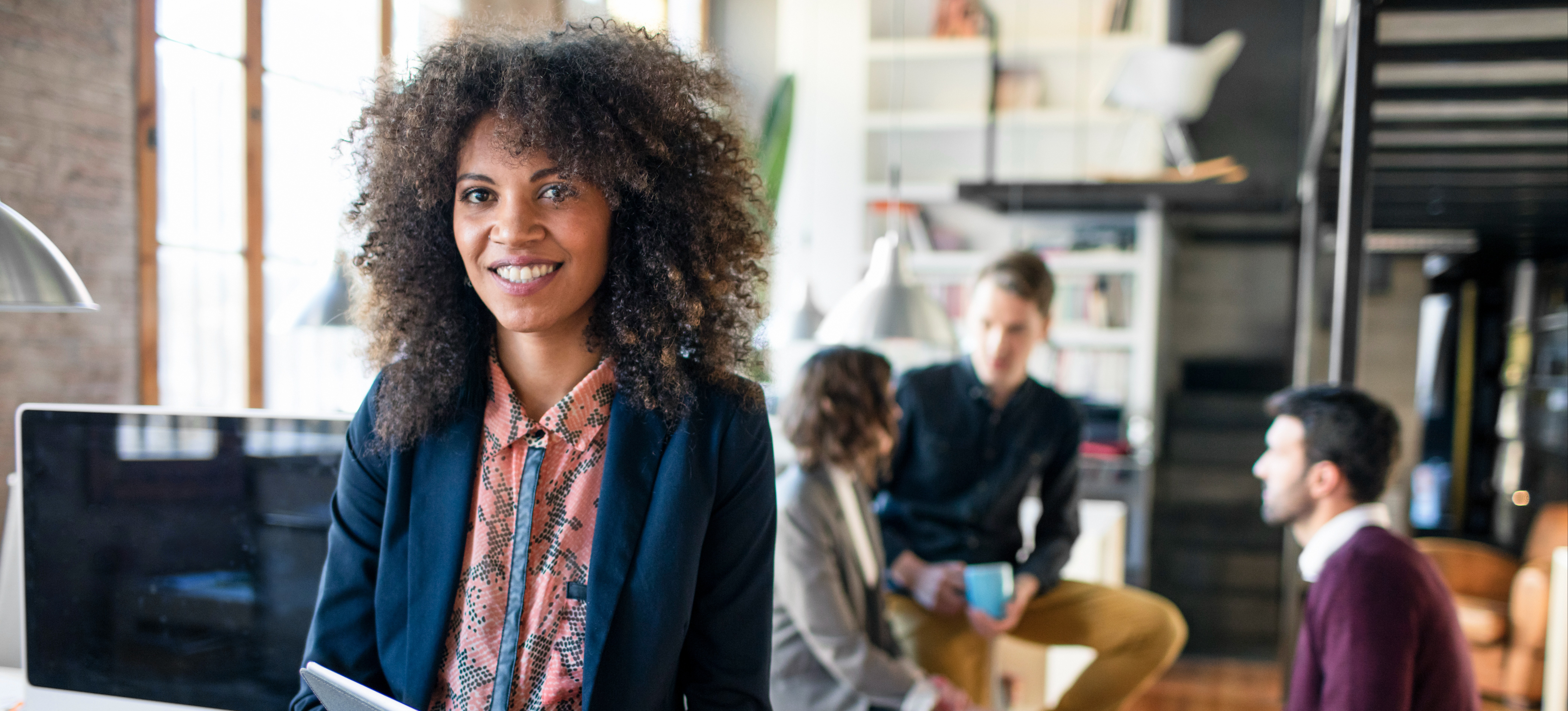 [Featured Image]:  Manager, wearing a blue jacket and printed top, sitting on a desk, preparing to communicate with team members. 