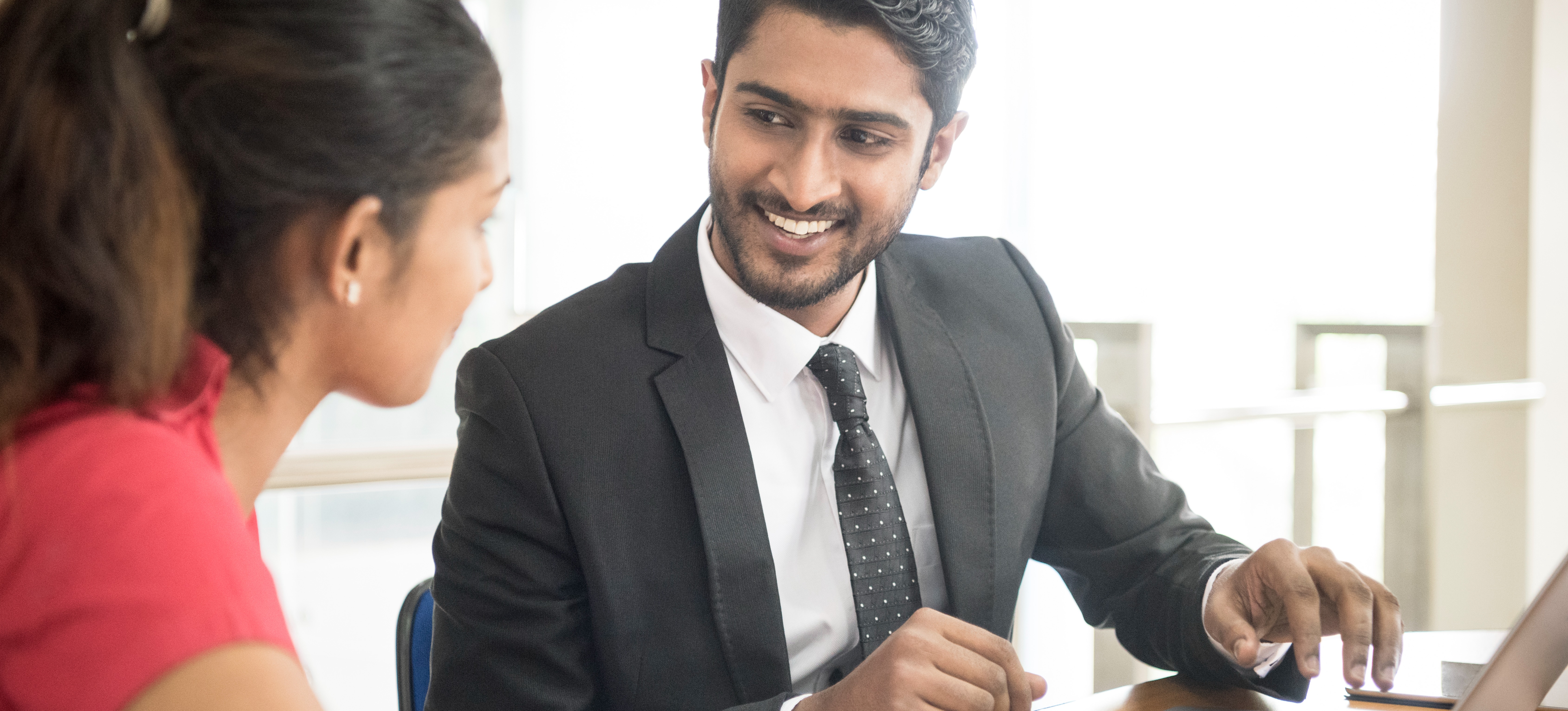 [Featured Image] A smiling tax accountant dressed in a suit and tie sits with their client at a table in a well-lit office and uses their laptop to explain to the client their finances.