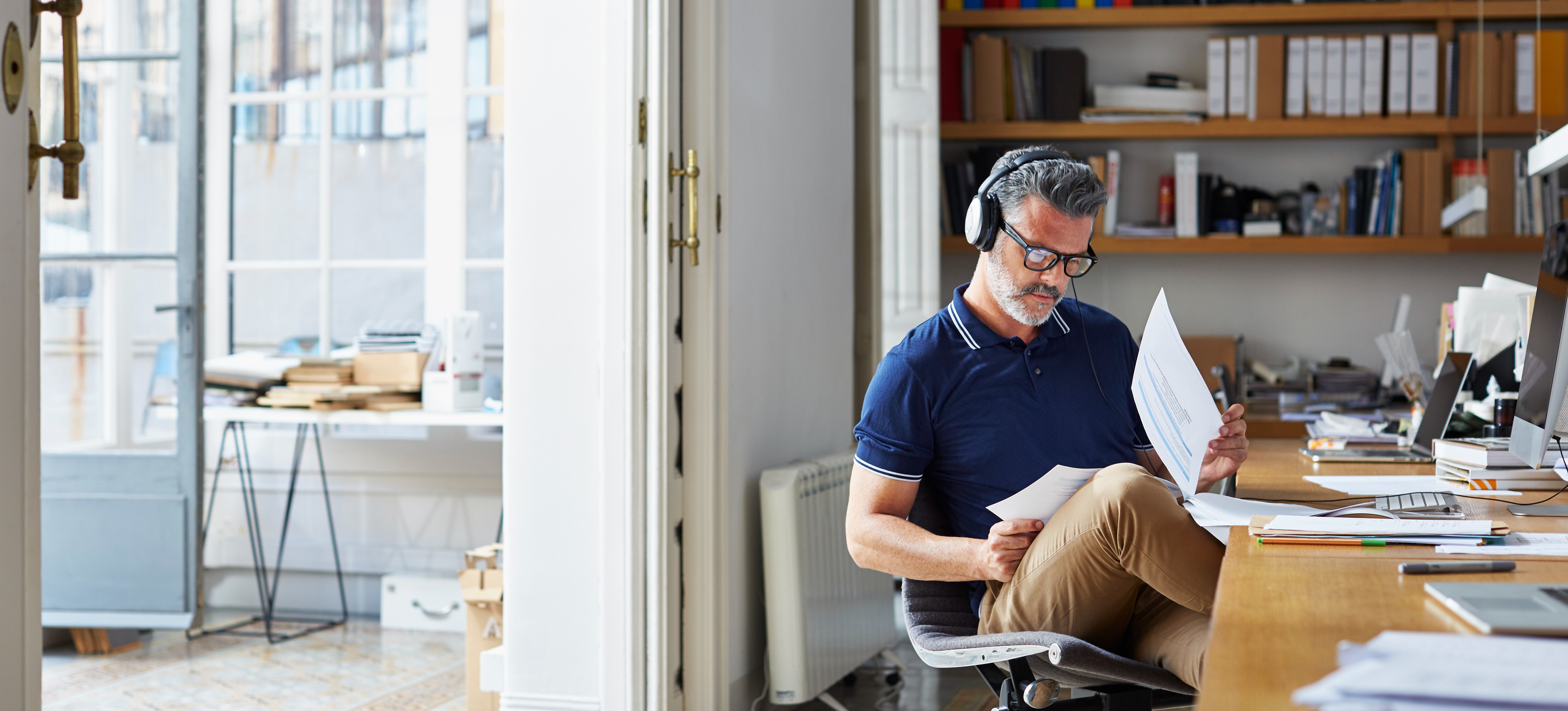 [Featured Image] A businessman working for a business intelligence analyst salary sits at his desk in a home office and analyzes business documents while wearing headphones.
