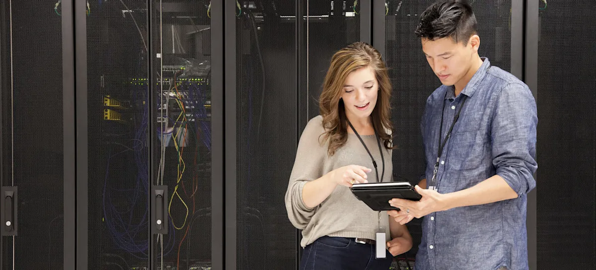 [Featured image] Two cybersecurity professionals wearing name badges and work-casual clothing stand in a server room and look at information on a tablet held by one of them.
