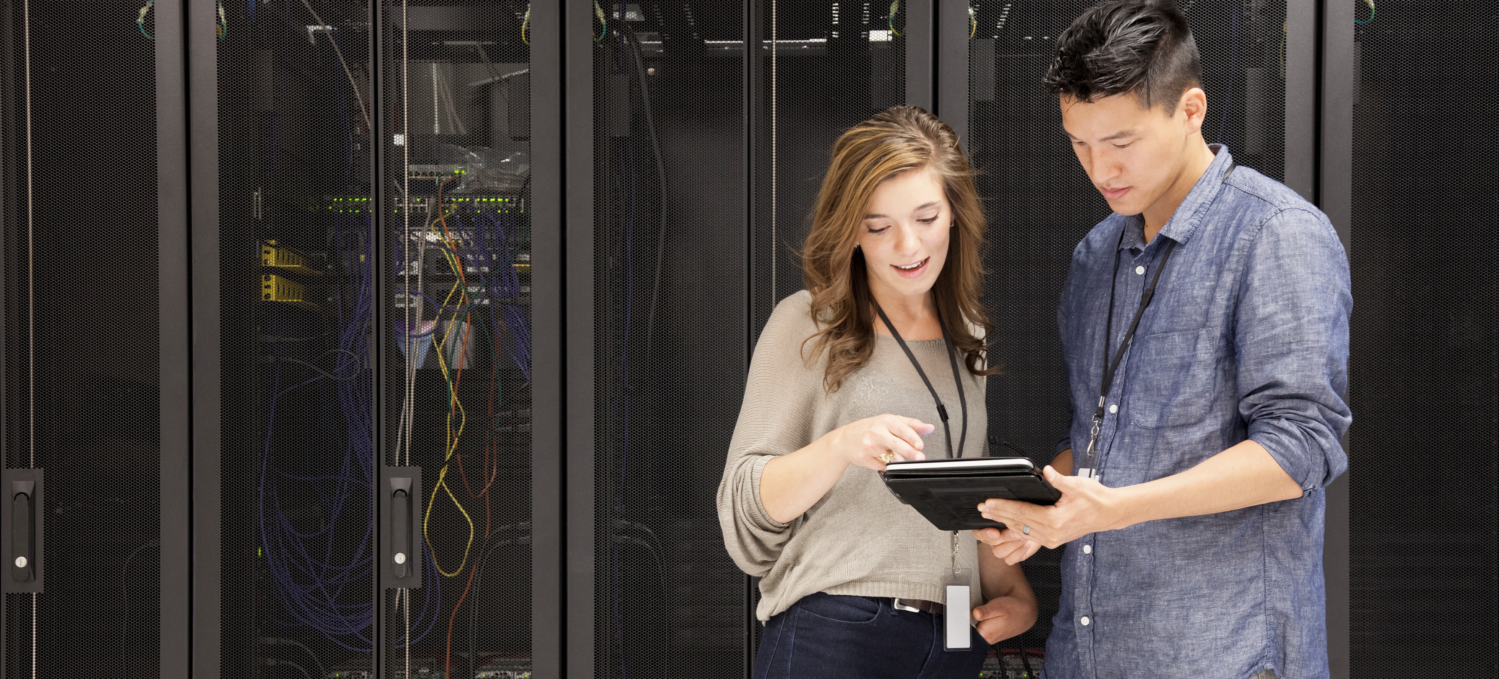 [Featured image] Two cybersecurity professionals wearing name badges and work-casual clothing stand in a server room and look at information on a tablet held by one of them.
