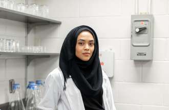 [Featured Image]:  A toxicologist, wearing a white lab coat, head covering and blue gloves is working in the laboratory. 