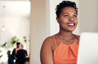 [Featured image] A young Black woman smiles off into the distance while sitting in front of her laptop.
