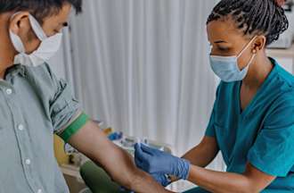 [Featured Image] A phlebotomist wearing scrubs draws blood from a patient in a hospital. 