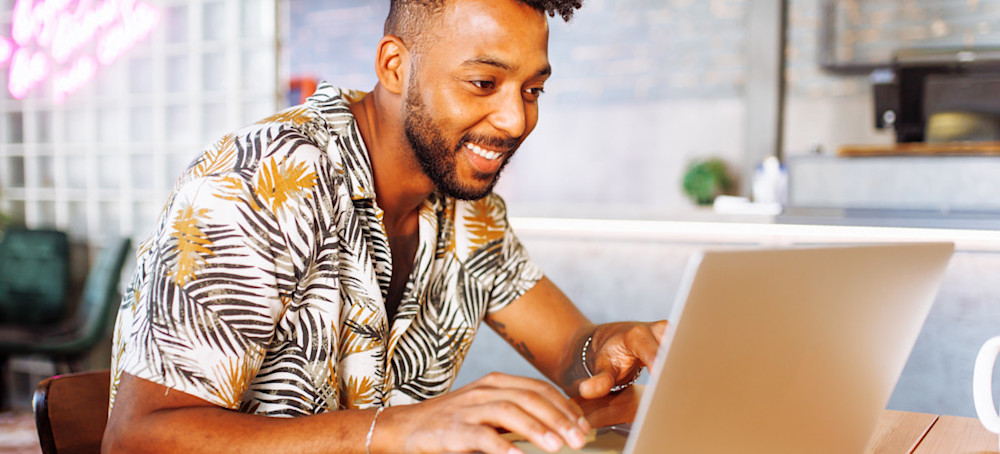 [Featured Image] A social media coordinator sits at a desk working on a campaign with his laptop and smartphone. 
