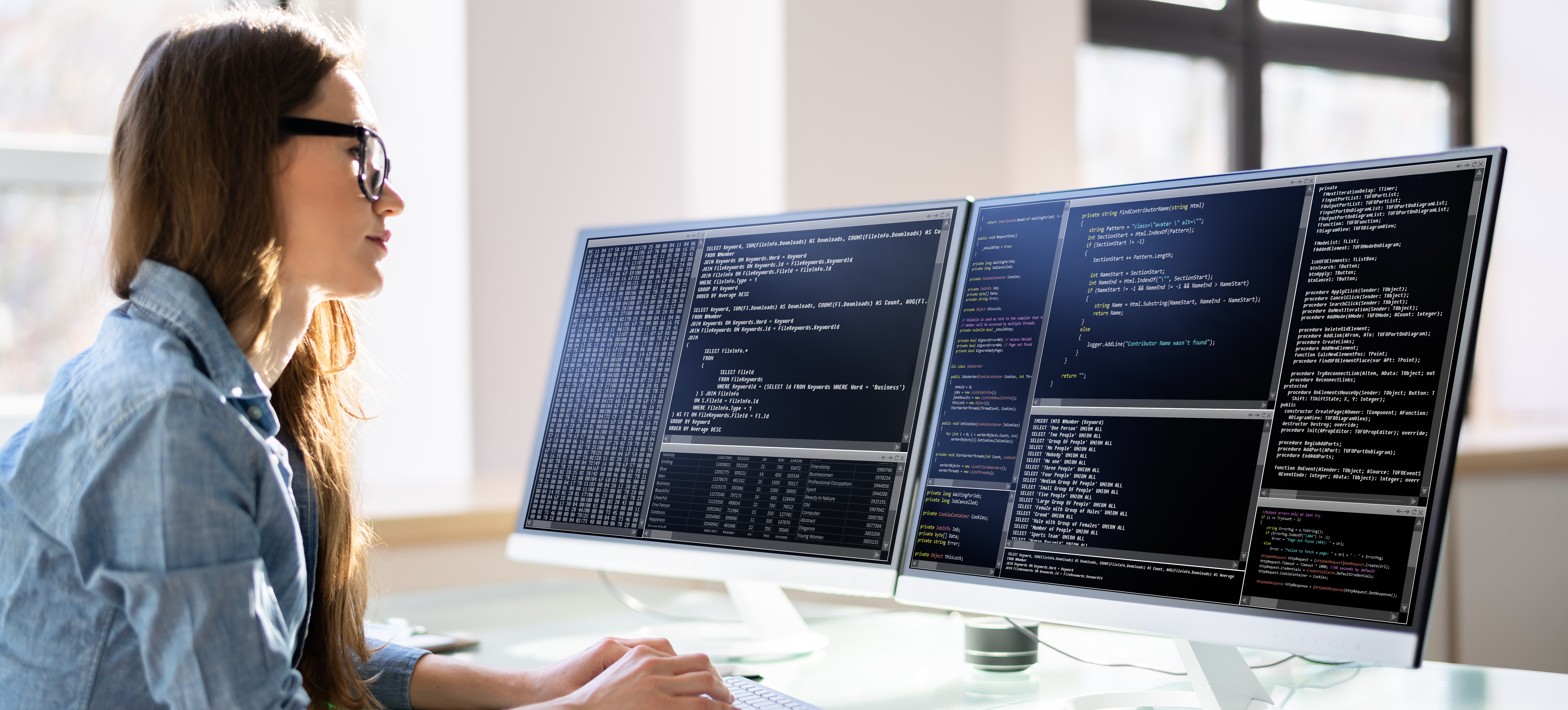 [Featured Image] A programmer, sits at her glass-top desk and works on two screens filled with code.