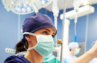 [Featured Image]:  A Licensed Vocational Nurse, wearing a purple uniform and a face covering, is looking at the vital signs of a patient. 