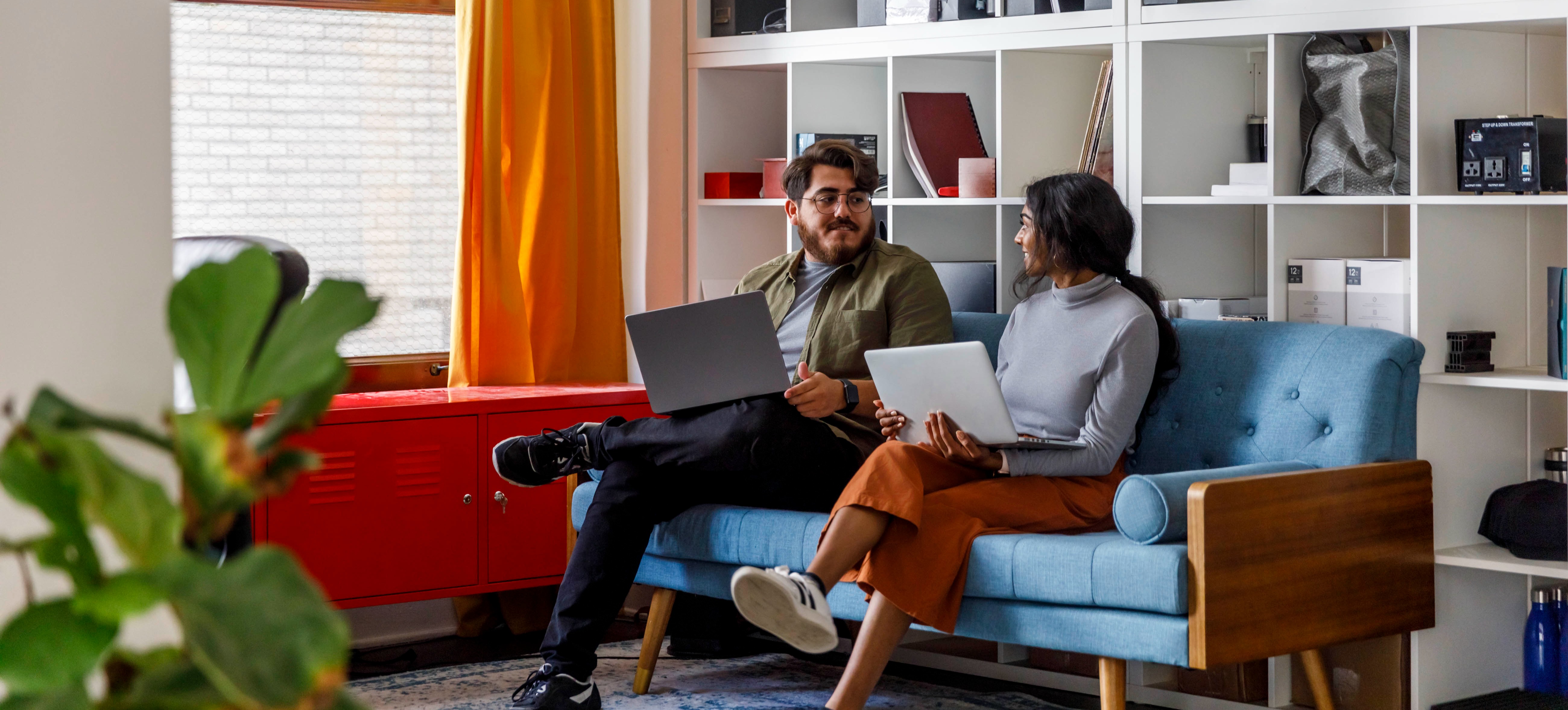 [Featured Image] Two learners sit on a blue couch in front of a large white bookshelf as they study probabilistic programming together and discuss its uses. 
