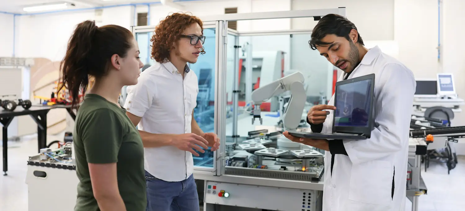 [Featured Image] A teacher in an engineering lab is showing two students how AI can help them make money.
