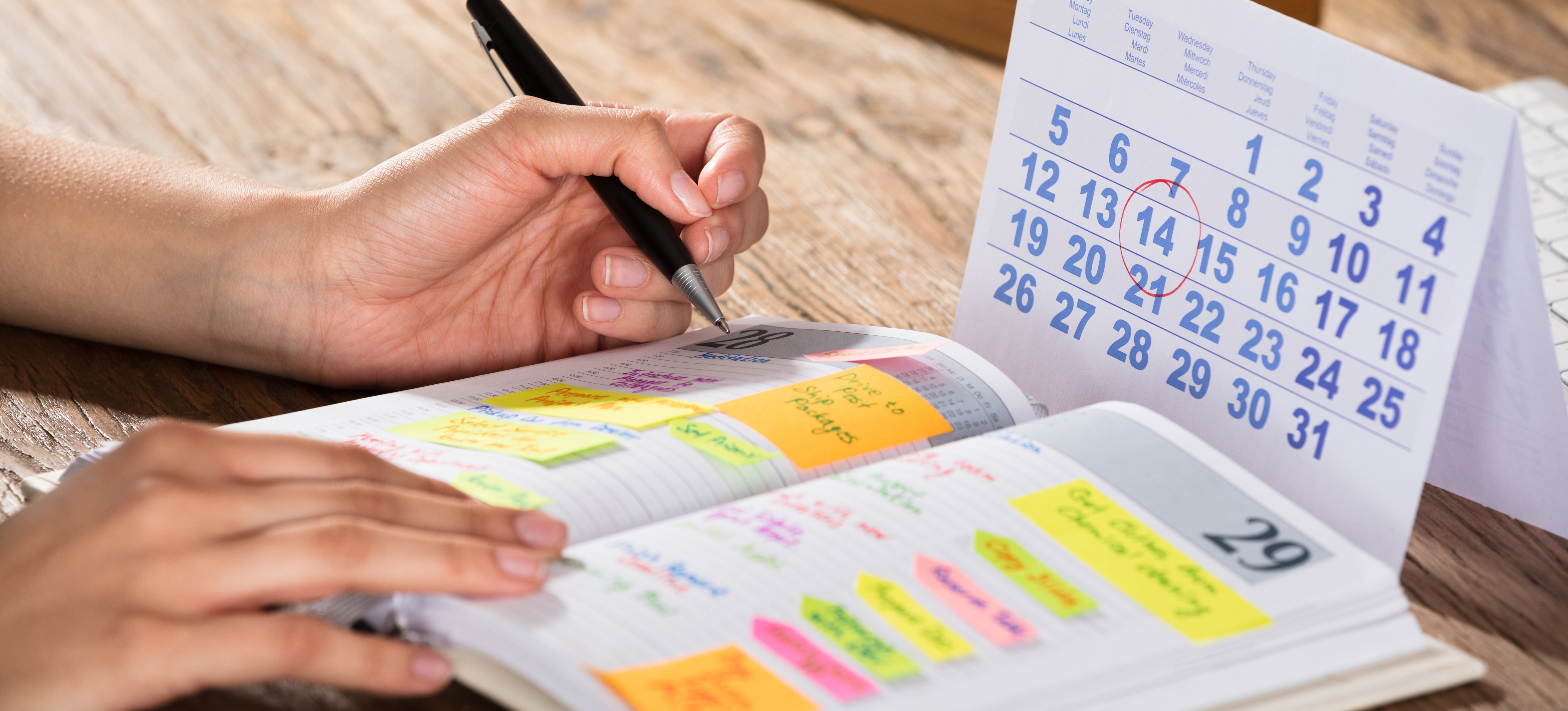 [Featured Image] A person holding a pen looks at the notes and highlights in their planner, as well as a calendar, as they consider how to prioritize tasks.