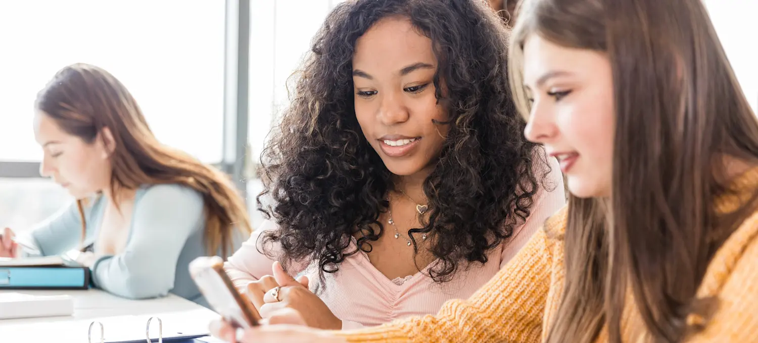 [Featured Image] Two students sit in a classroom and look at a cell phone to determine when college applications are due.
