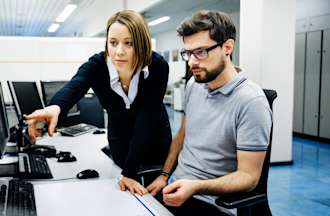 [Featured Image] A person in a black sweater points out an item on a computer screen to a coworker in glasses seated in front of their desktop computer.