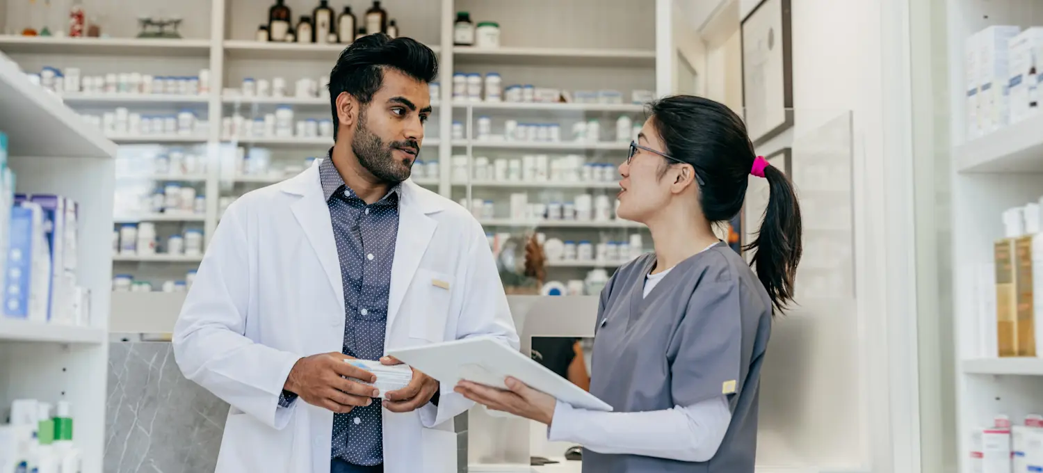 [Featured Image] Two pharmacists stand in a pharmacy and discuss the need for cybersecurity in the pharma industry. 
