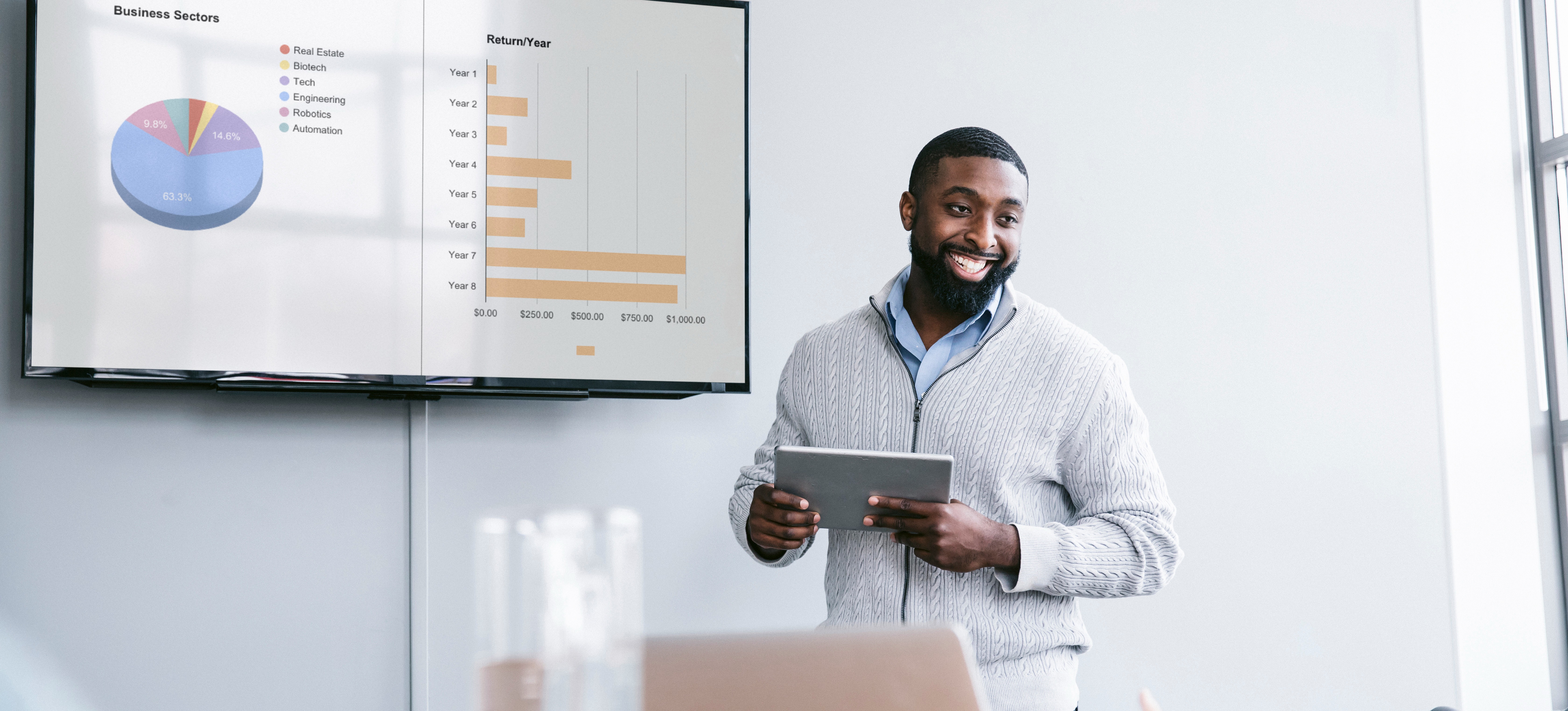 [Featured Image] A man who understands data definition presents a data set to his colleagues, standing in front of a presentation on a screen and while holding a tablet and looking out at the conference room.
