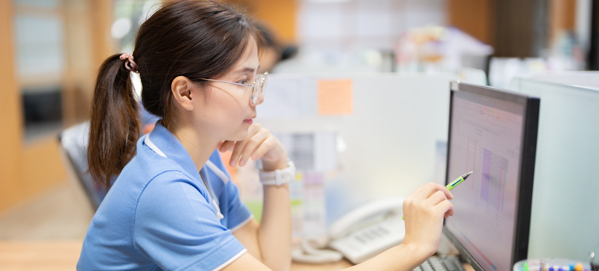 [Featured Image] A medical coder working in an office works on her computer while consulting a large handbook of medical codes.
