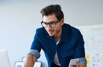 [Featured Image]:  A male, wearing a blue shirt and glasses, is working on his laptop and holding his cellphone, as he works on his development project.