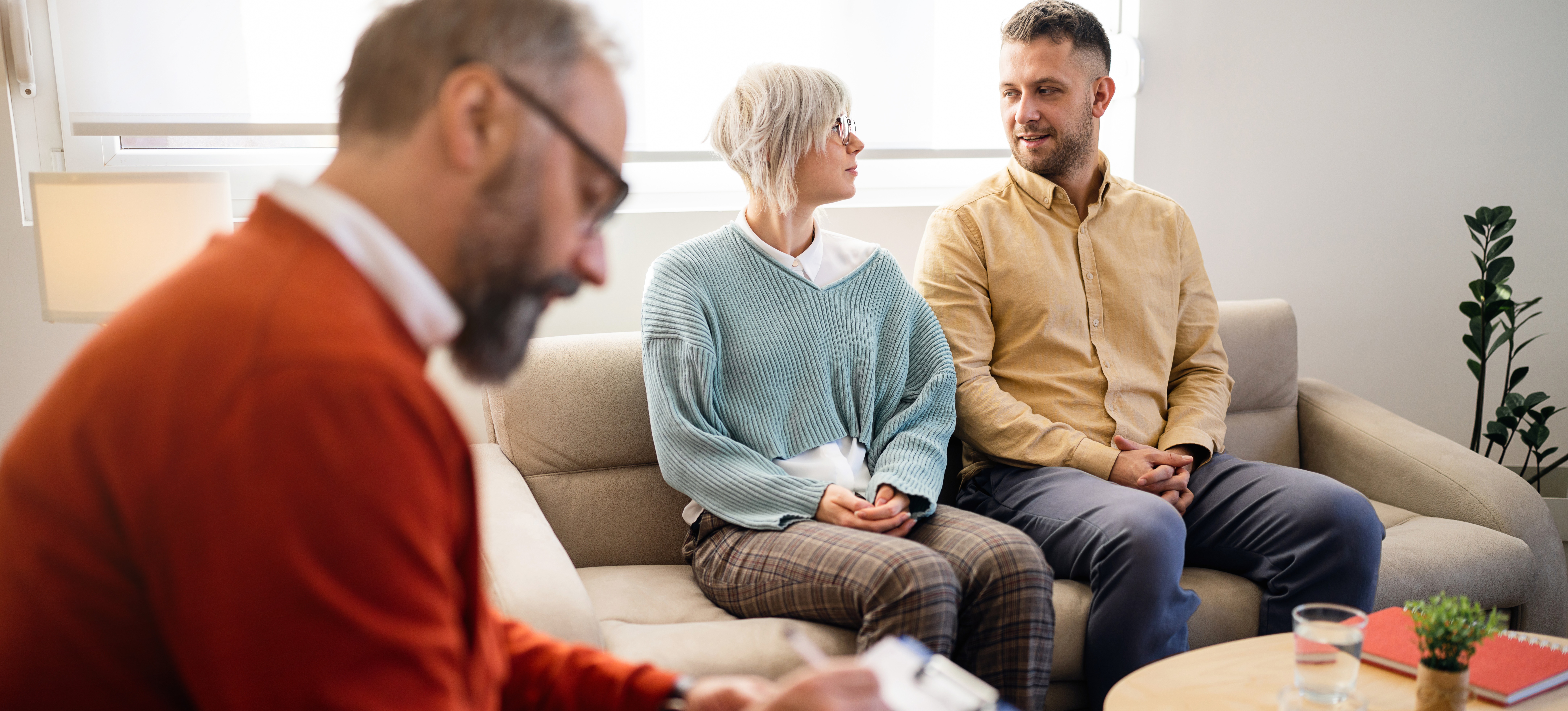[Featured Image] A man and woman sit on a sofa in a therapist's office as the counselor takes notes, demonstrating a career you can pursue after earning a master's in psychology.
