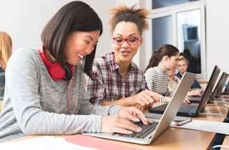 [Featured image] Two women practice writing Python code on their laptops in a classroom lab setting.