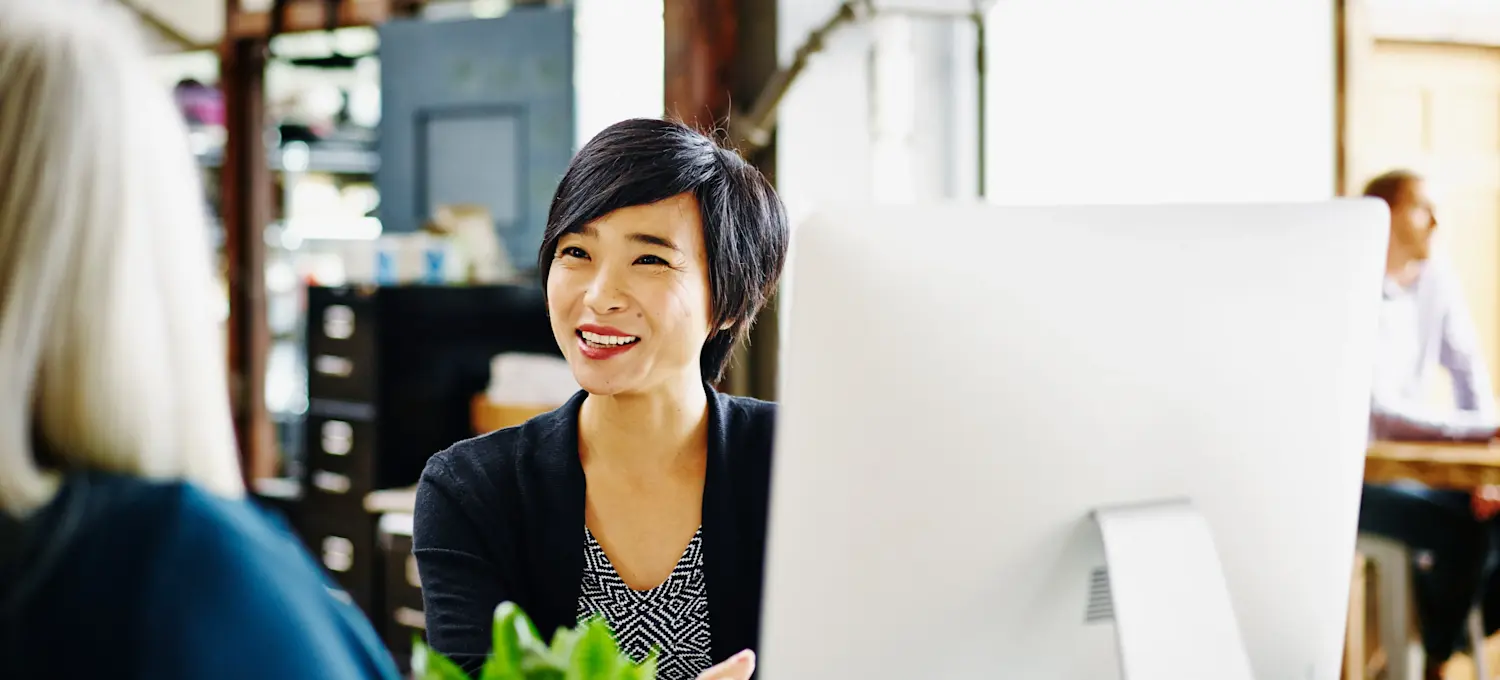 [Featured Image] A program manager sits at their computer in an office setting and talks with their colleague about the various programs their team is involved in.
