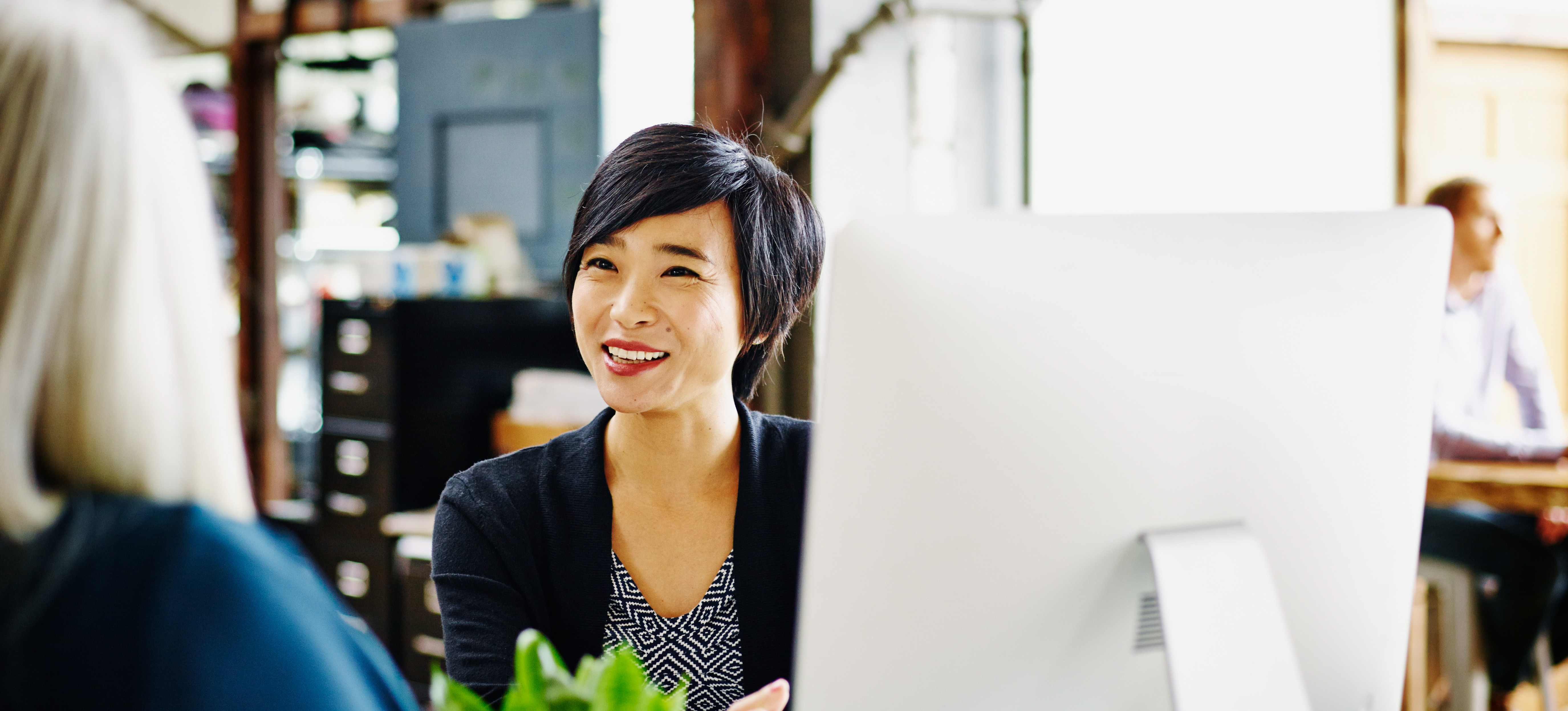 [Featured Image] A program manager sits at her computer in an office setting and talks with her colleague about the various programs their team is involved in.
