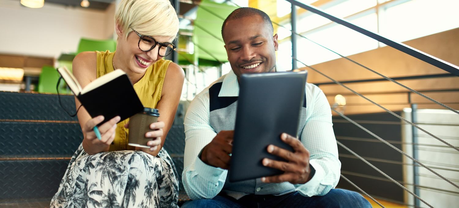 [Featured Image] Two smiling social media specialists sit on steps in an office and discuss their marketing strategy.