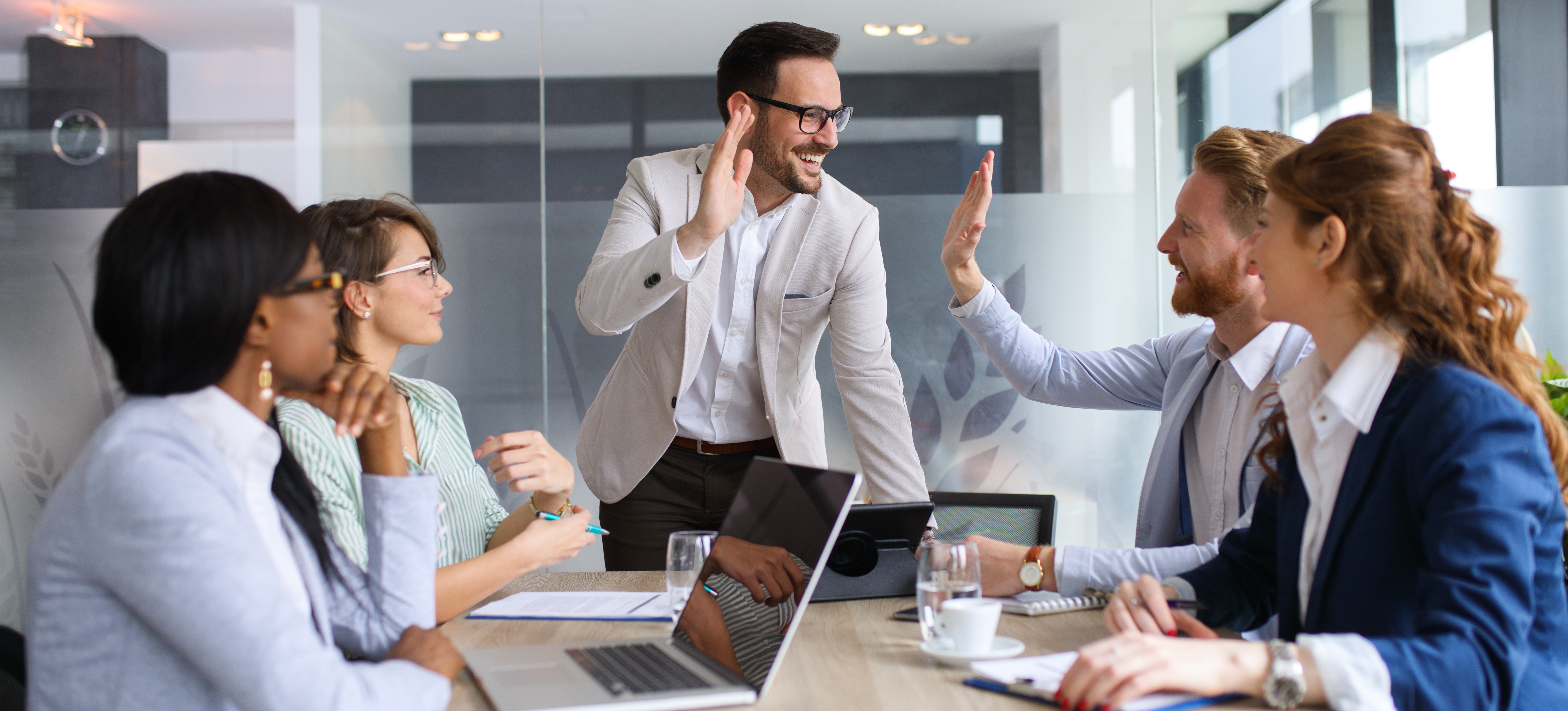 [Featured Image] A sales manager high fives a colleague during a team meeting with three other salespeople. 
