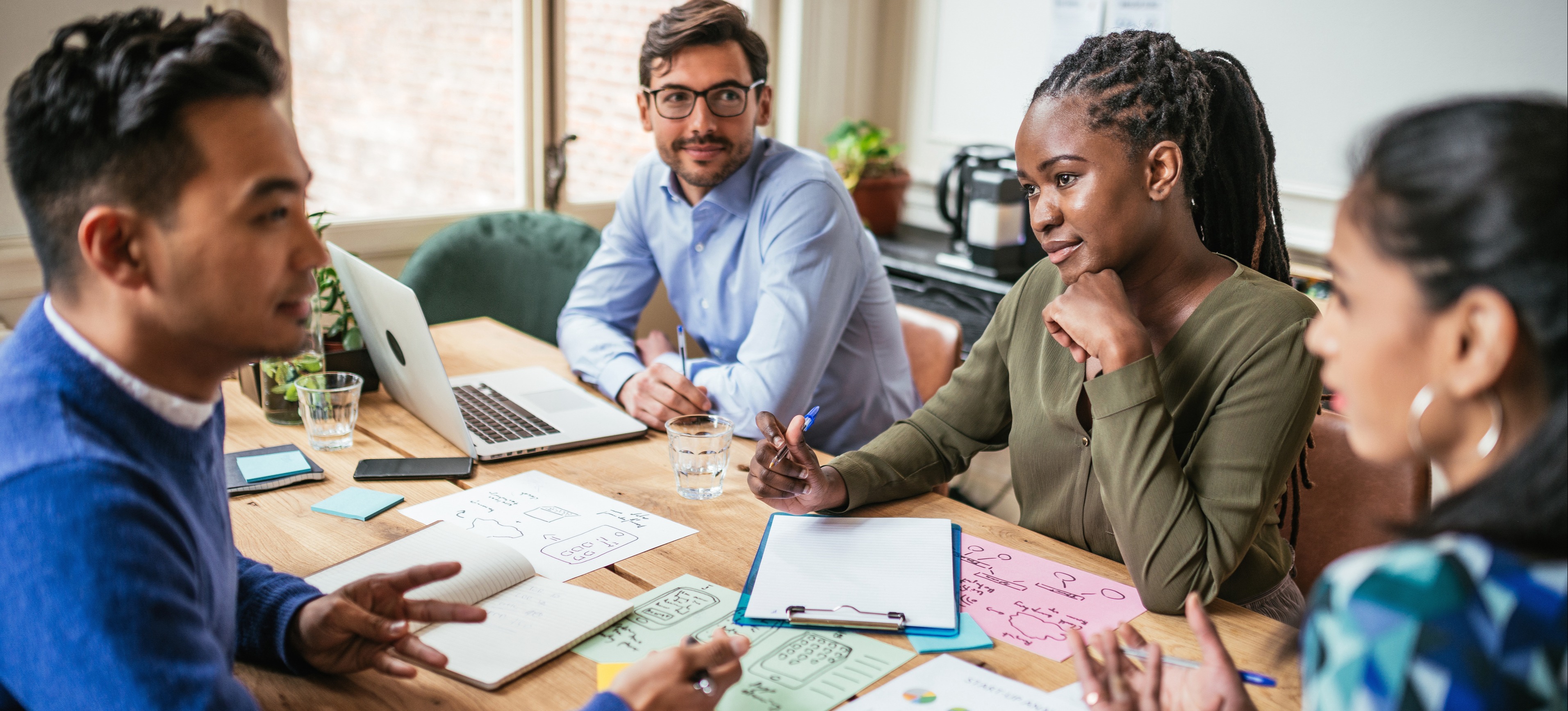 [Featured image] Four people sit around a conference table, scattered with papers and charts, and have a conversation. 