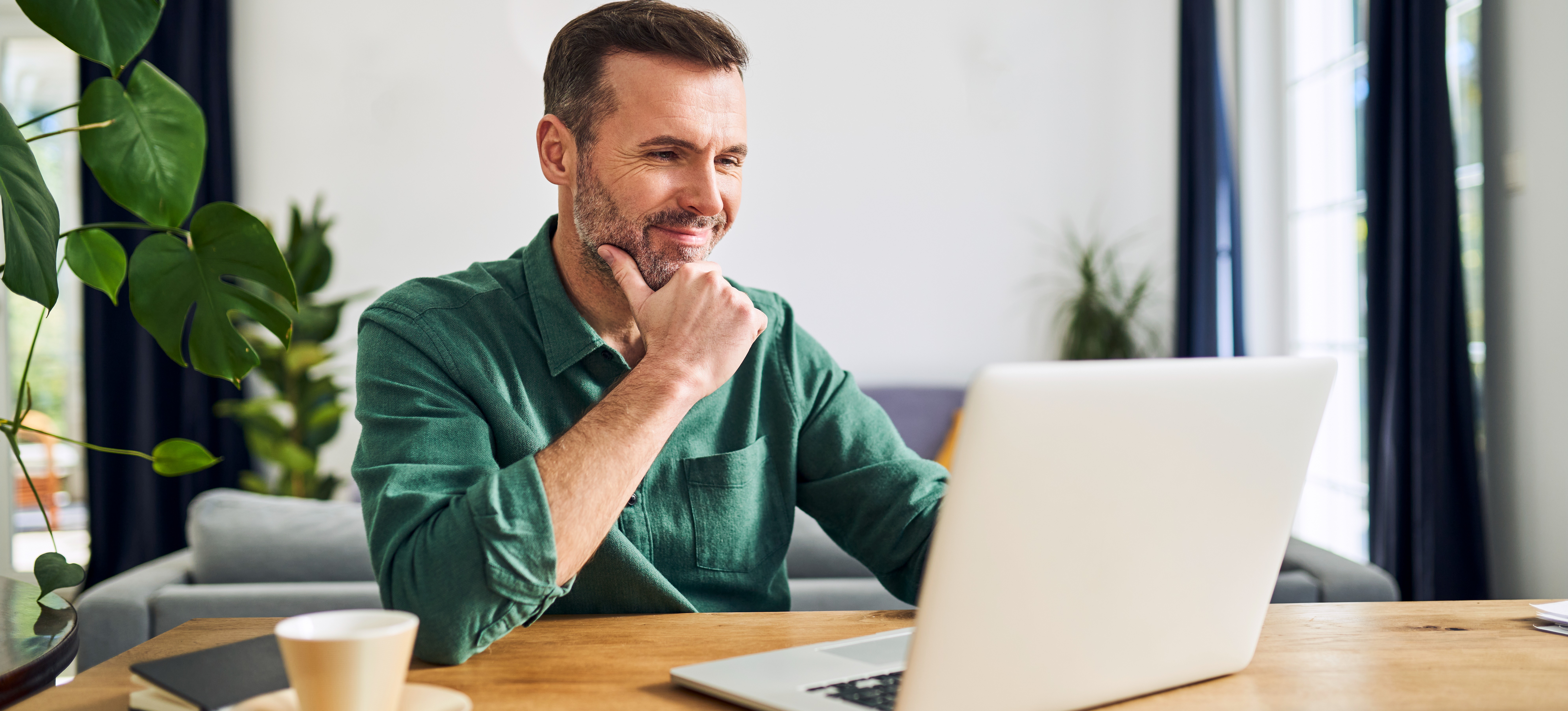 [Featured Image] A man working from home on a laptop considers several LinkedIn headline examples before choosing his own.