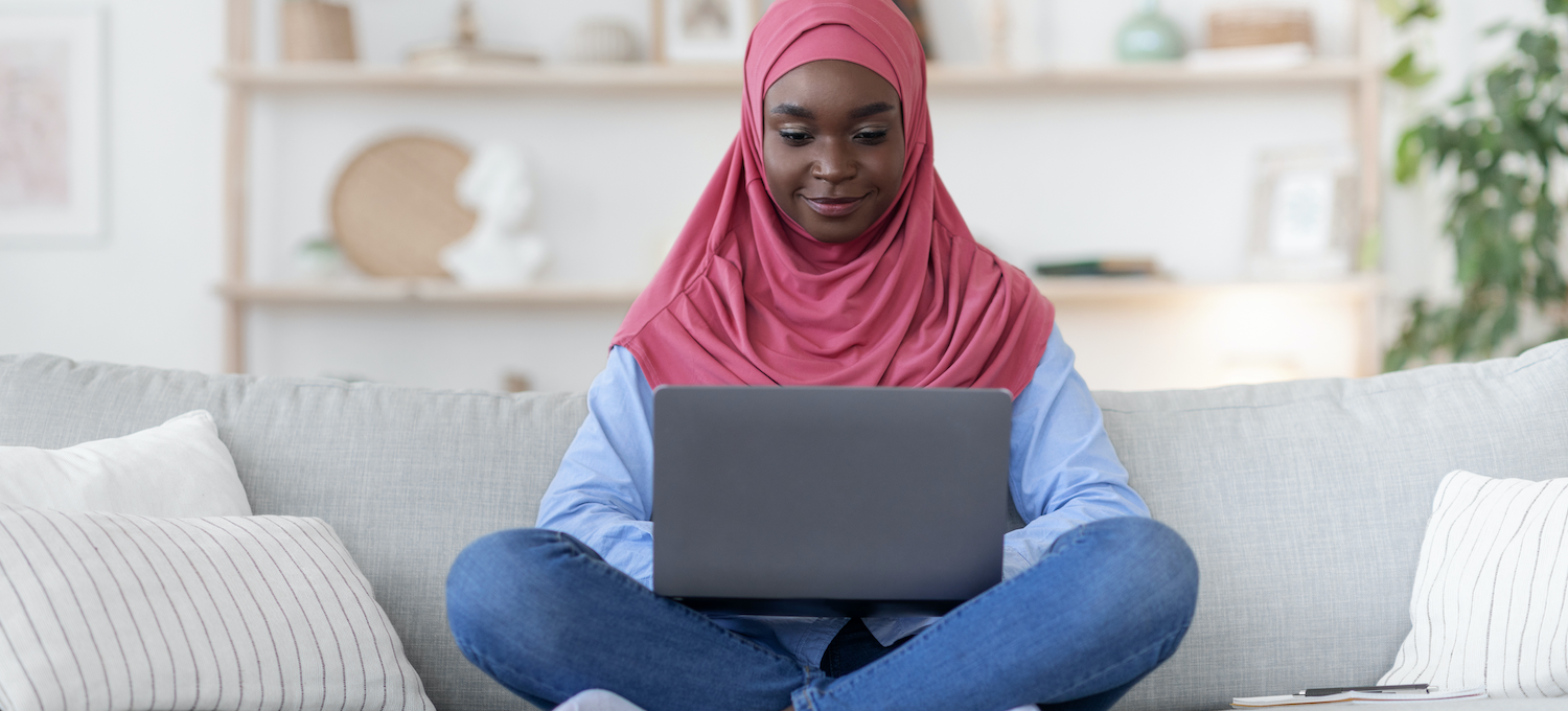 [Featured image] A smiling person wearing a pink al-amira sits on a grey sofa working on a laptop computer.