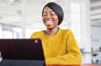 [Featured image] Job candidate smiles at her tablet as she logs on for a video interview with a hiring manager in an open office space.