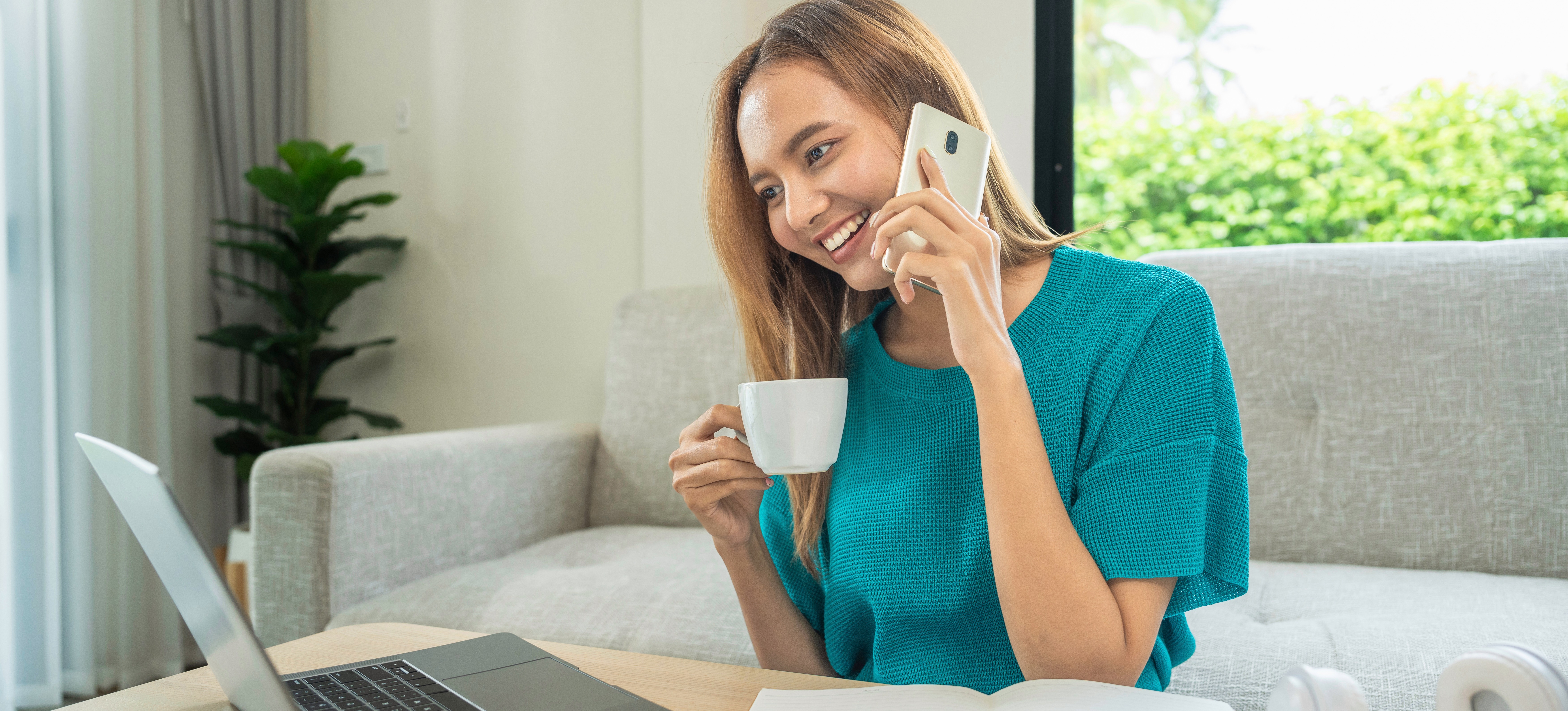[Featured Image] A woman sits at home, talks on the phone, and uses her laptop while contemplating a career change after learning about a prompt engineering salary. 
