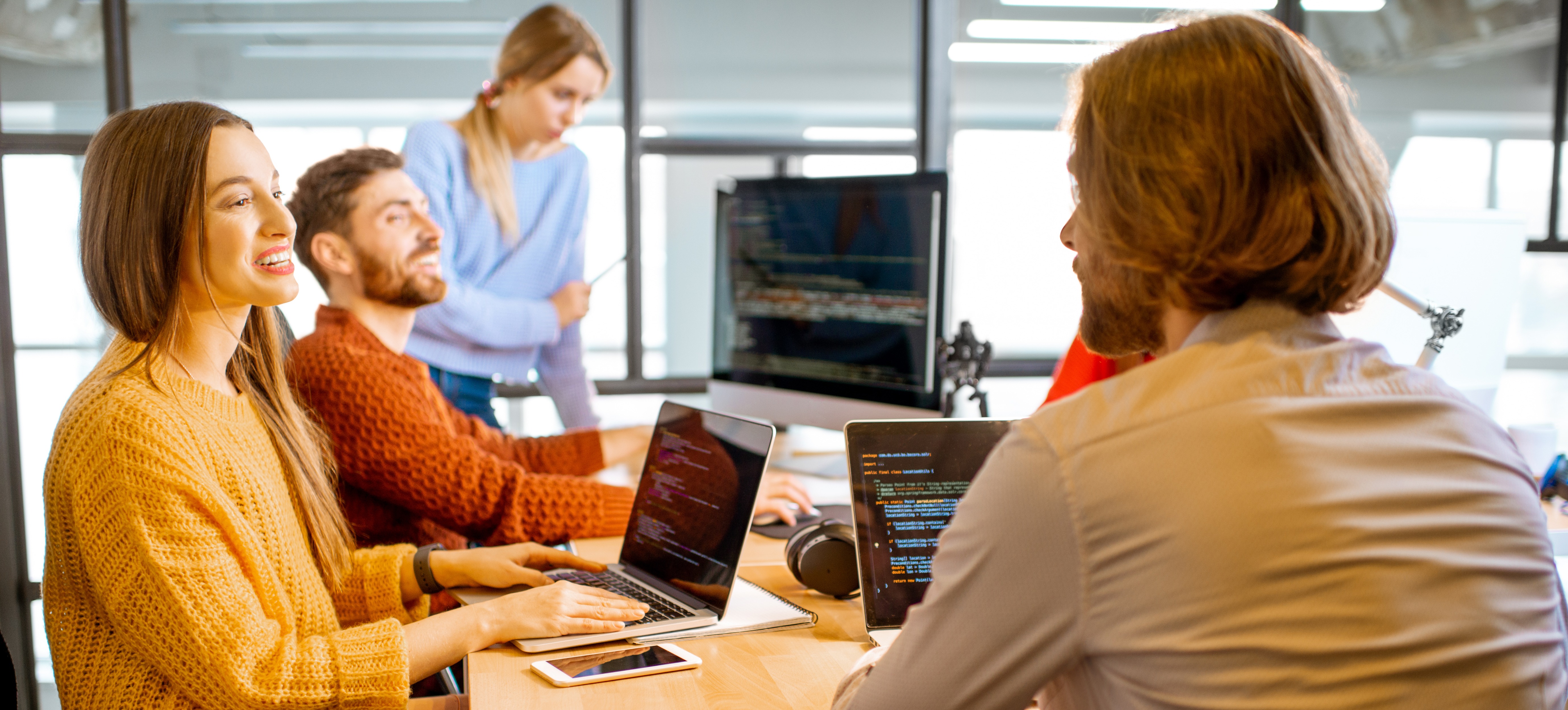 [Featured Image] A happy group of engineers working on their laptops at a communal table debate the benefits of a DevOps engineer vs. cloud engineer career path.
