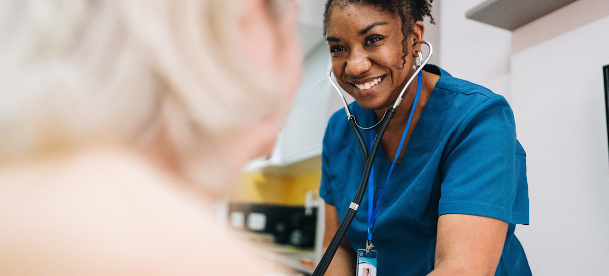 [Featured Image] A health care worker, who receives a community health worker salary, visits an elderly person in their home to provide health services like taking blood pressure using a stethoscope.

