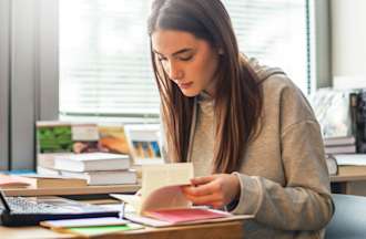 [Featured Image] A woman is sitting at her desk looking through a book with a laptop in front of her.