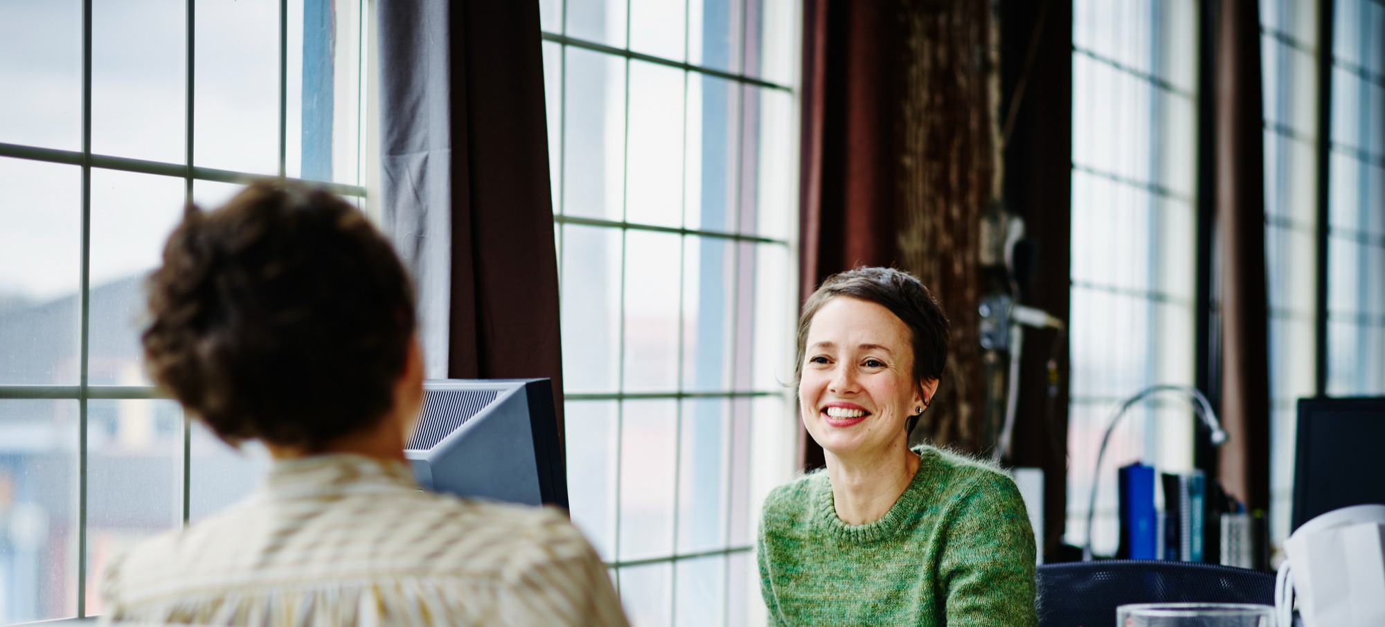 [Featured image] A female, wearing a green top, is sitting at her desk by the window in the office, as she performs her duties as an hr coordinator. 