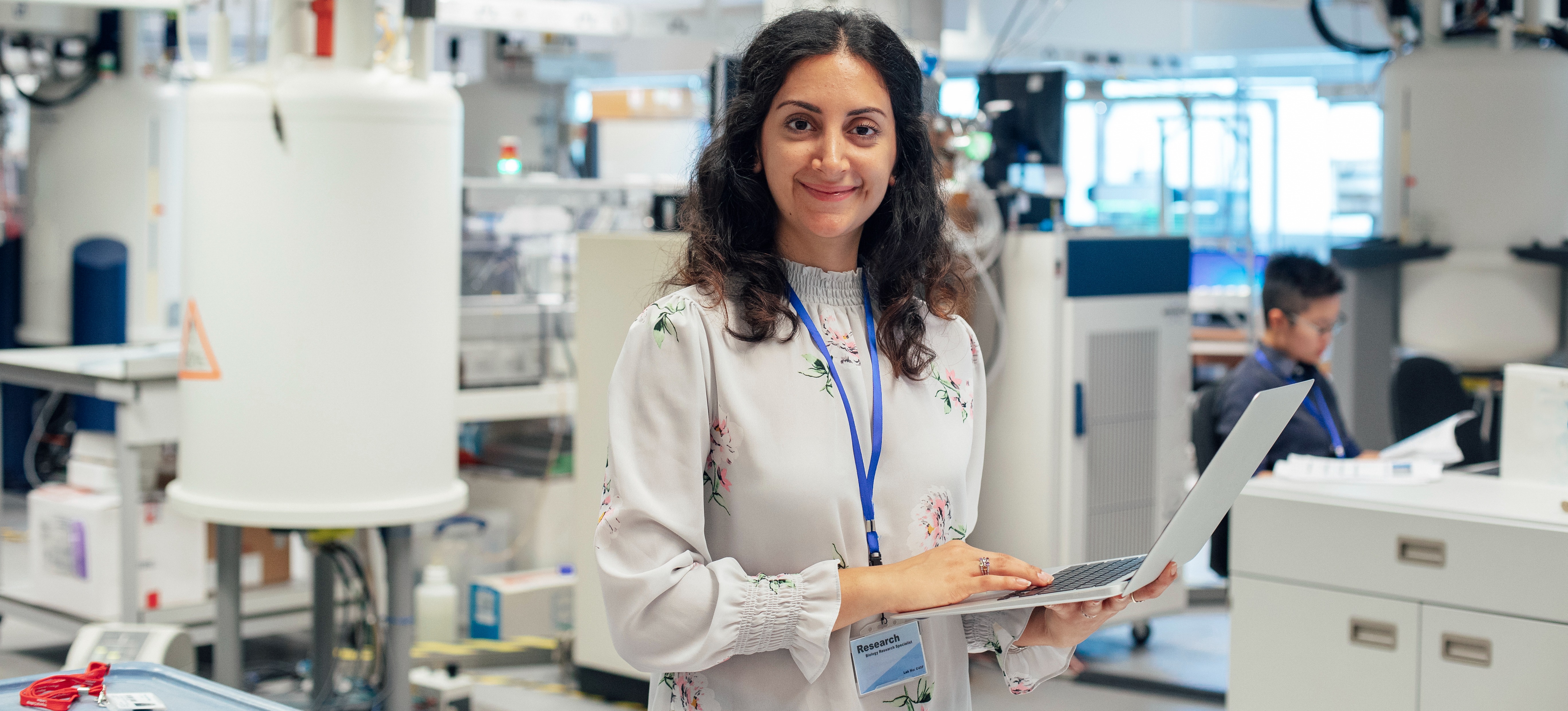 [Featured image] Pharmacist in white standing in front of medical equipment with a laptop computer