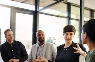 [Featured Image] Four members of the operations management team, two men and two women are meeting around the conference table. 