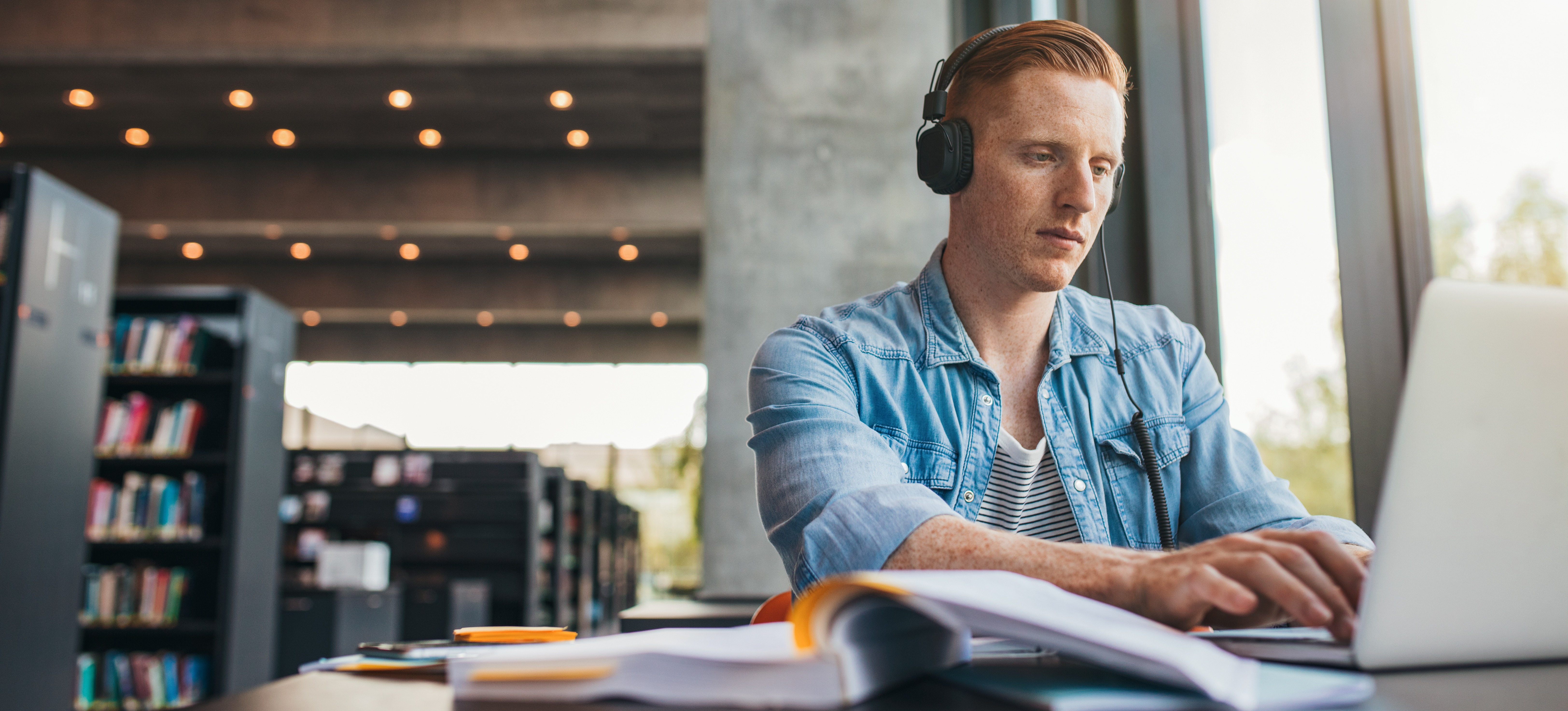[Featured image] A university student sits in a library studying with an open book and a laptop computer.