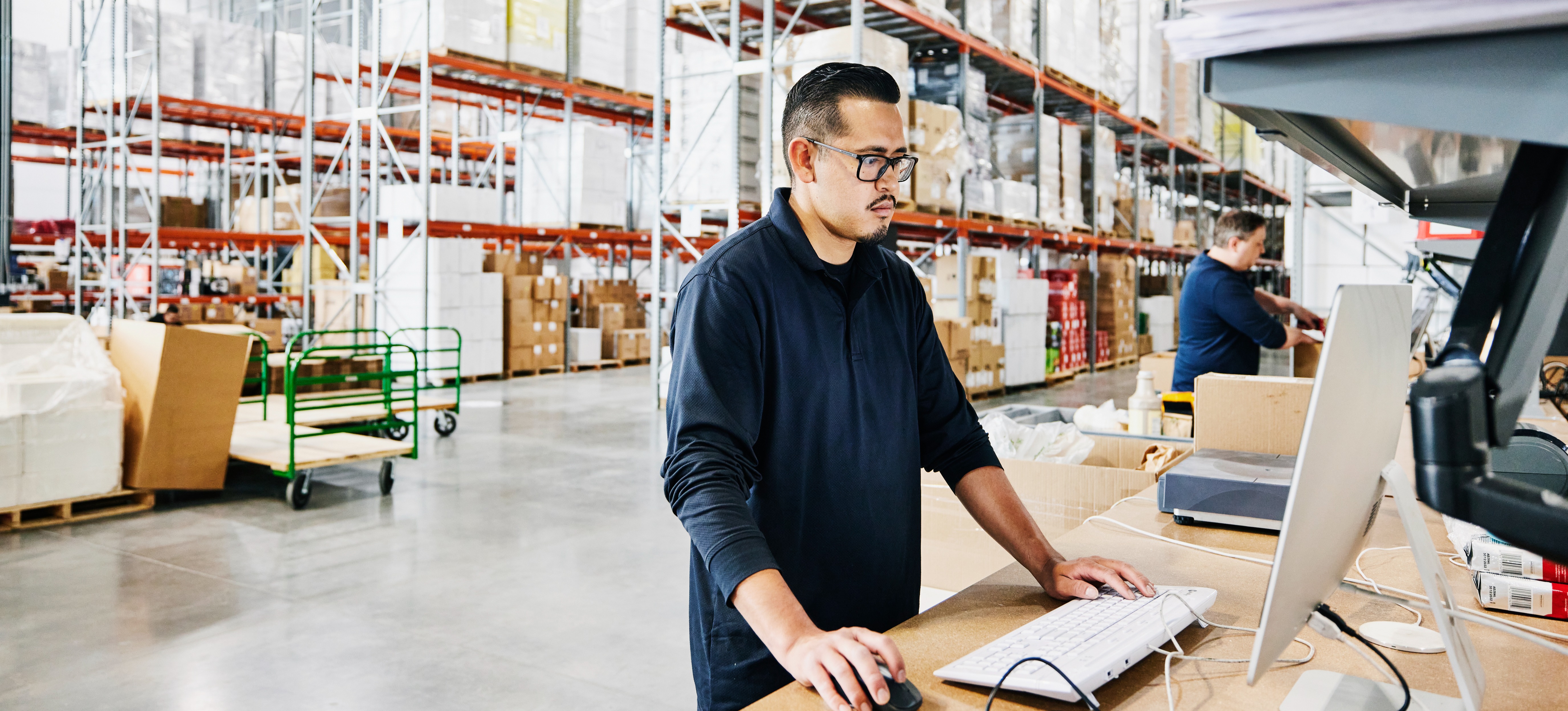 [Featured Image] A logistician stands at a table in a large warehouse and works on his computer.
