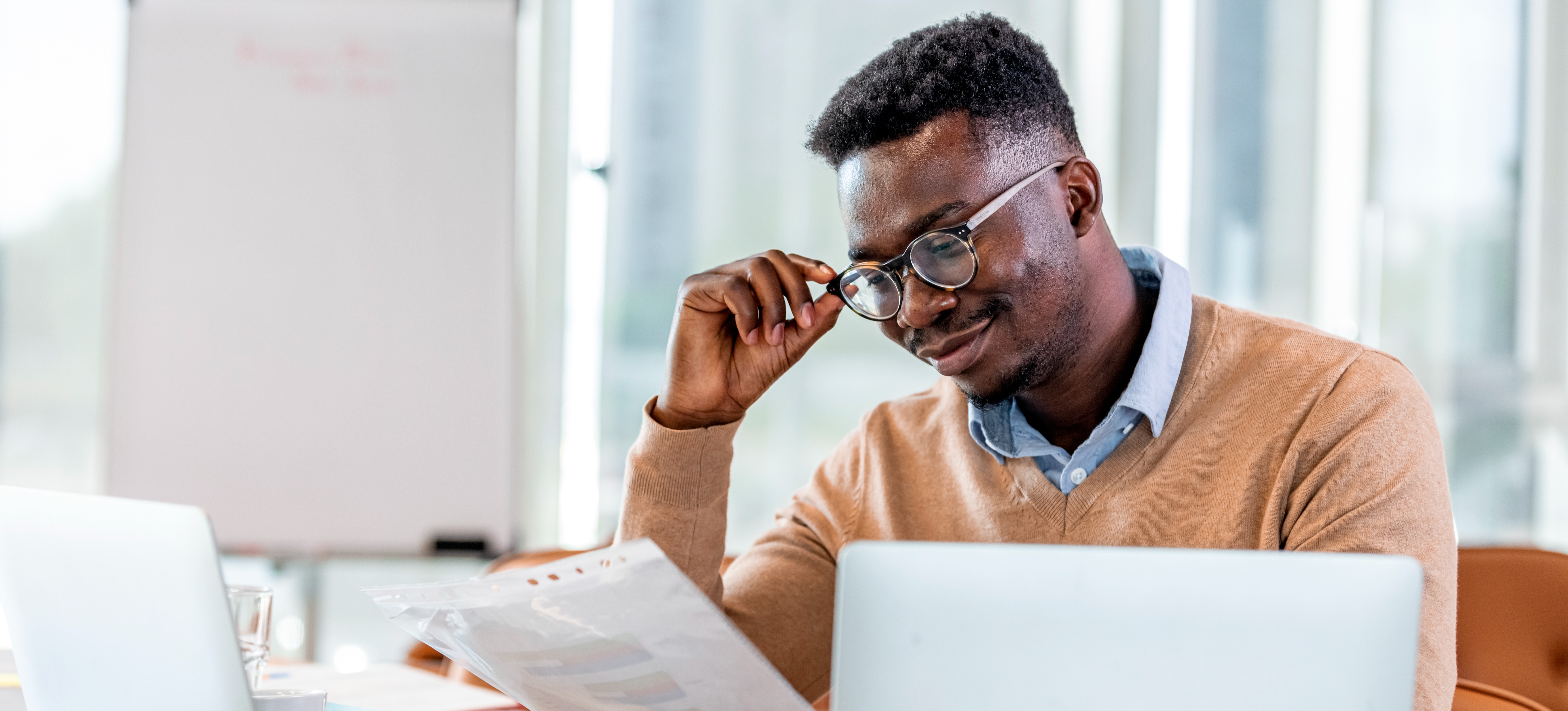 [Featured Image] A smiling financial controller tilts their glasses as they read a report that they hold in their other hand and sits in front of two computers in their office.
