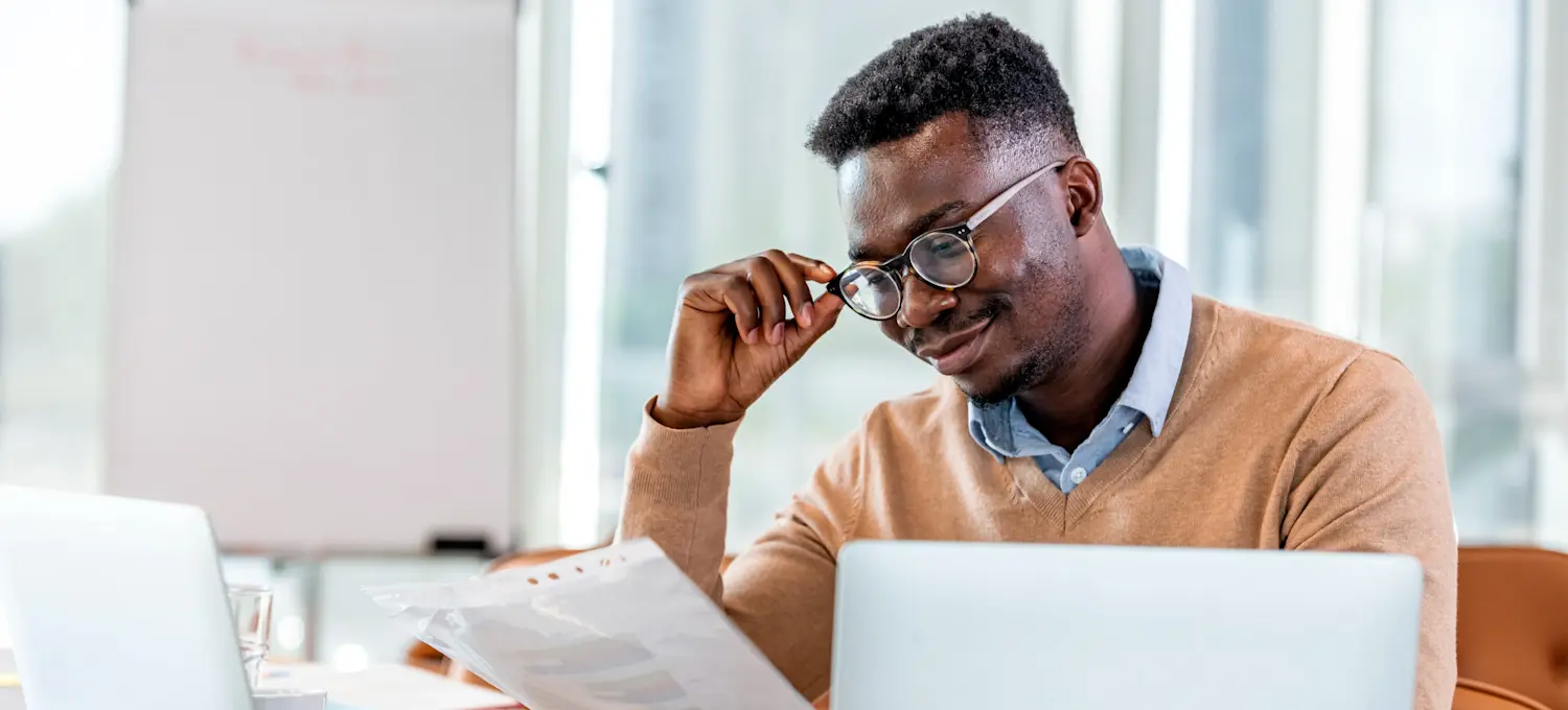[Featured Image] A smiling financial controller tilts their glasses as they read a report that they hold in their other hand and sits in front of two computers in their office.

