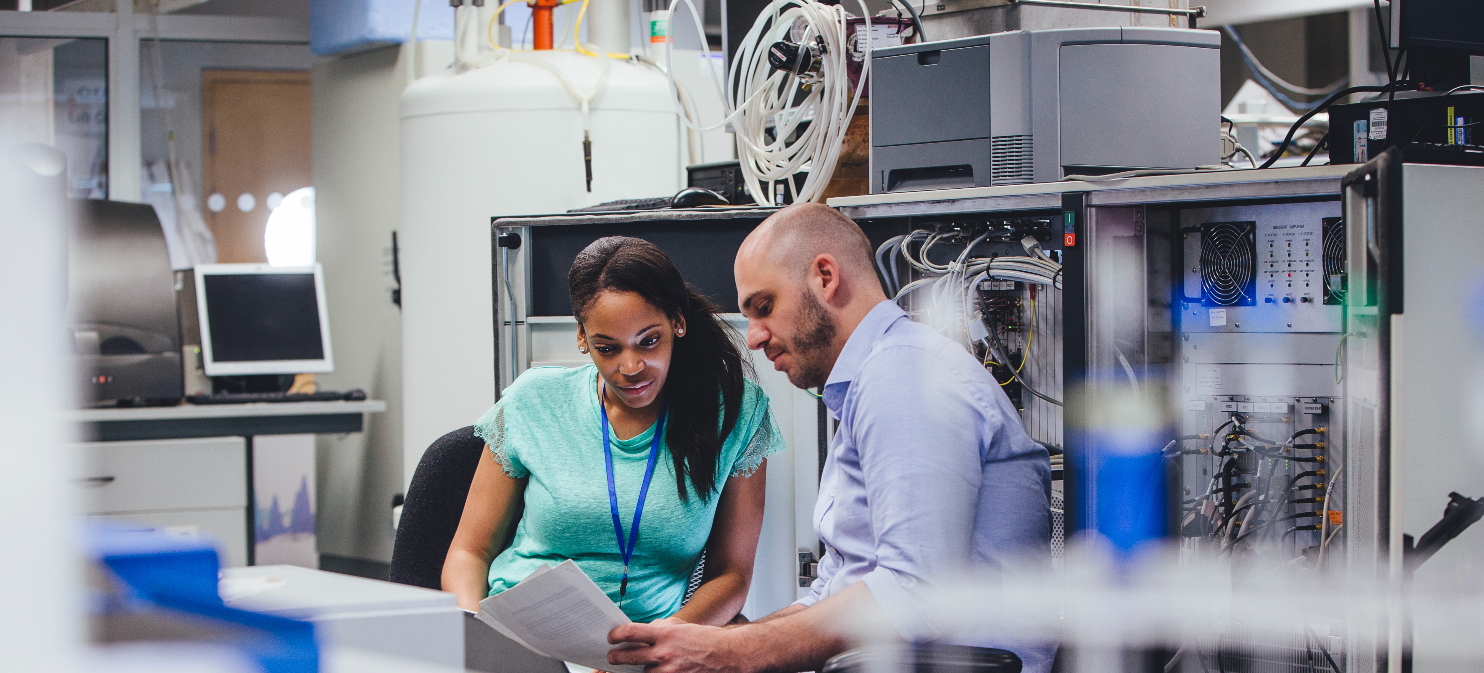 [Featured Image]:  Two professionals are discussing records and charts in a room with computers and big data information.